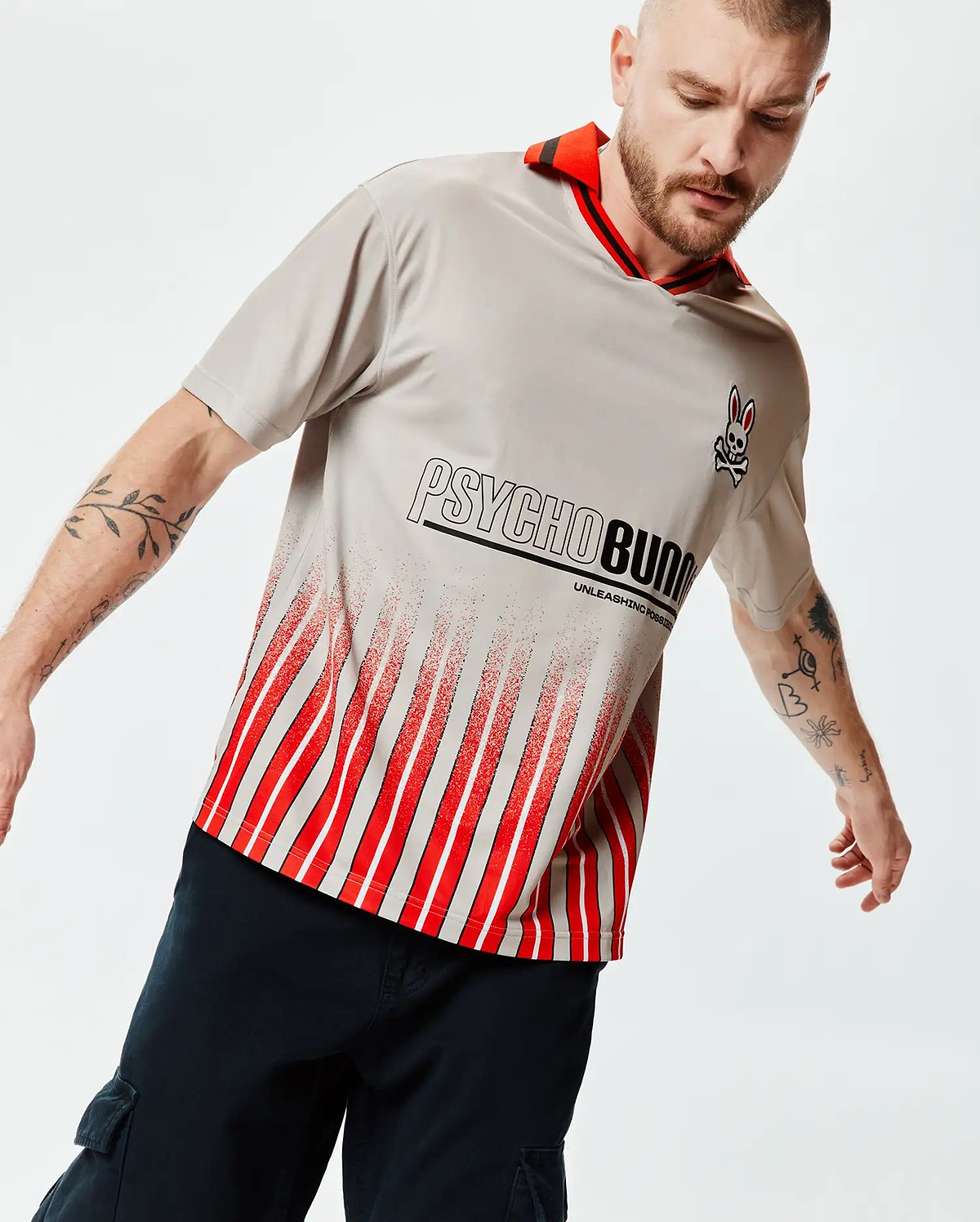 Mens Camden Oversized Soccer Jersey, light grey jersey knit, red striped collar, small Psycho Bunny skull logo, large Psycho Bunny text with Unleashing Possibilities, and a distressed red and white vertical stripe pattern. (3)