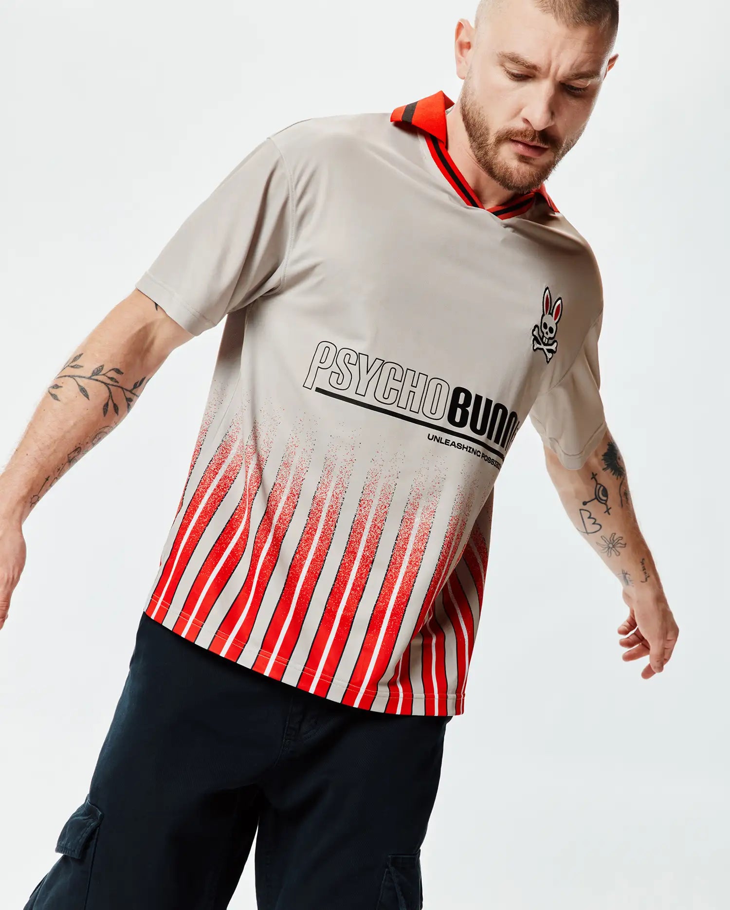 Mens Camden Oversized Soccer Jersey, light grey jersey knit, red striped collar, small Psycho Bunny skull logo, large Psycho Bunny text with Unleashing Possibilities, and a distressed red and white vertical stripe pattern. (3)