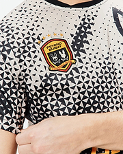 Mens Joshua Soccer Tee in off-white jersey with a black geometric triangle pattern, featuring a large embroidered Psycho Bunny Football Club patch with bunny skull and gold stars on the chest. (2)