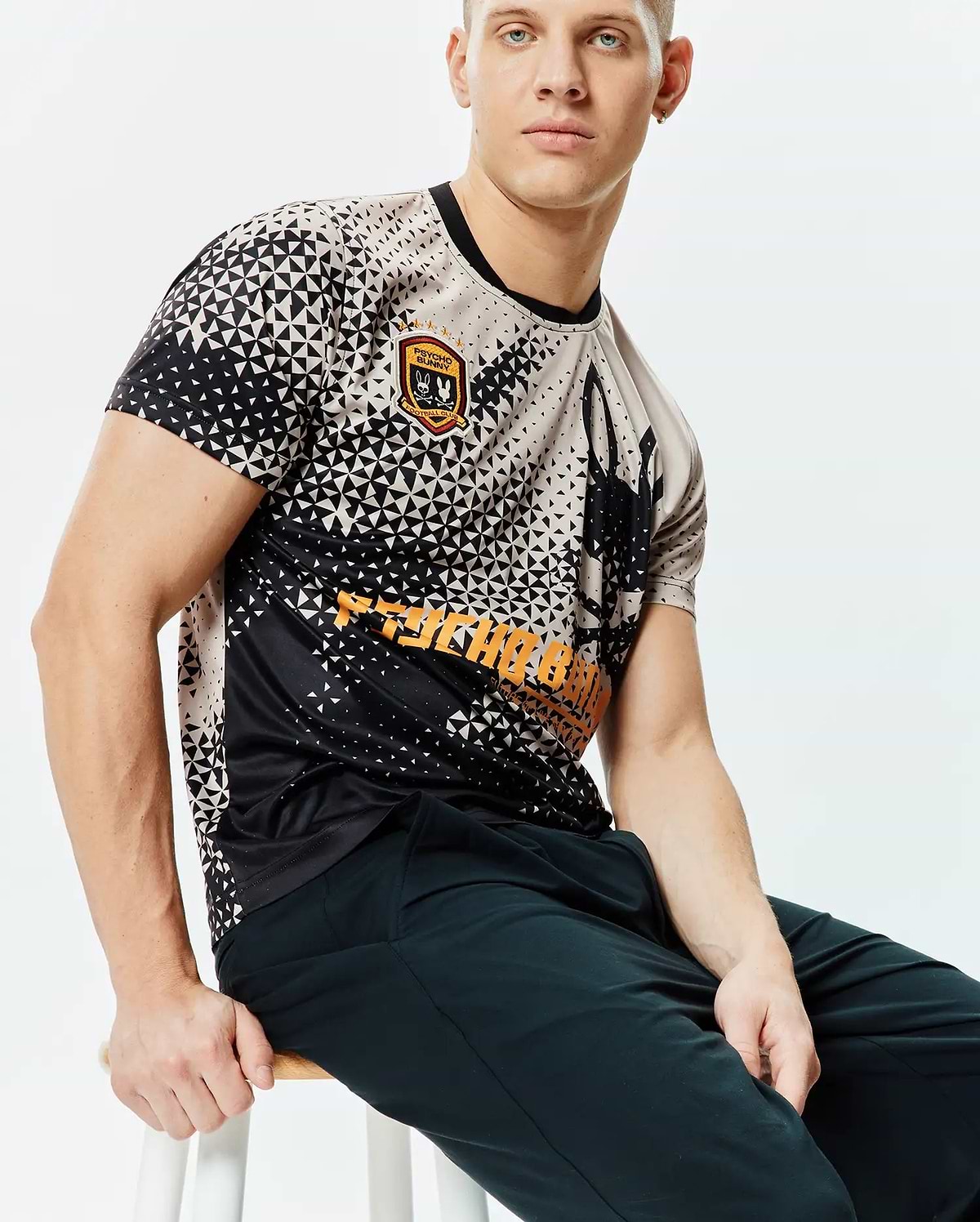 Mens Joshua Soccer Tee, a classic fit black and beige geometric triangle pattern jersey with an embroidered Psycho Bunny Football Club patch, large bunny skull graphic, and bold orange Psycho Bunny text. (3)