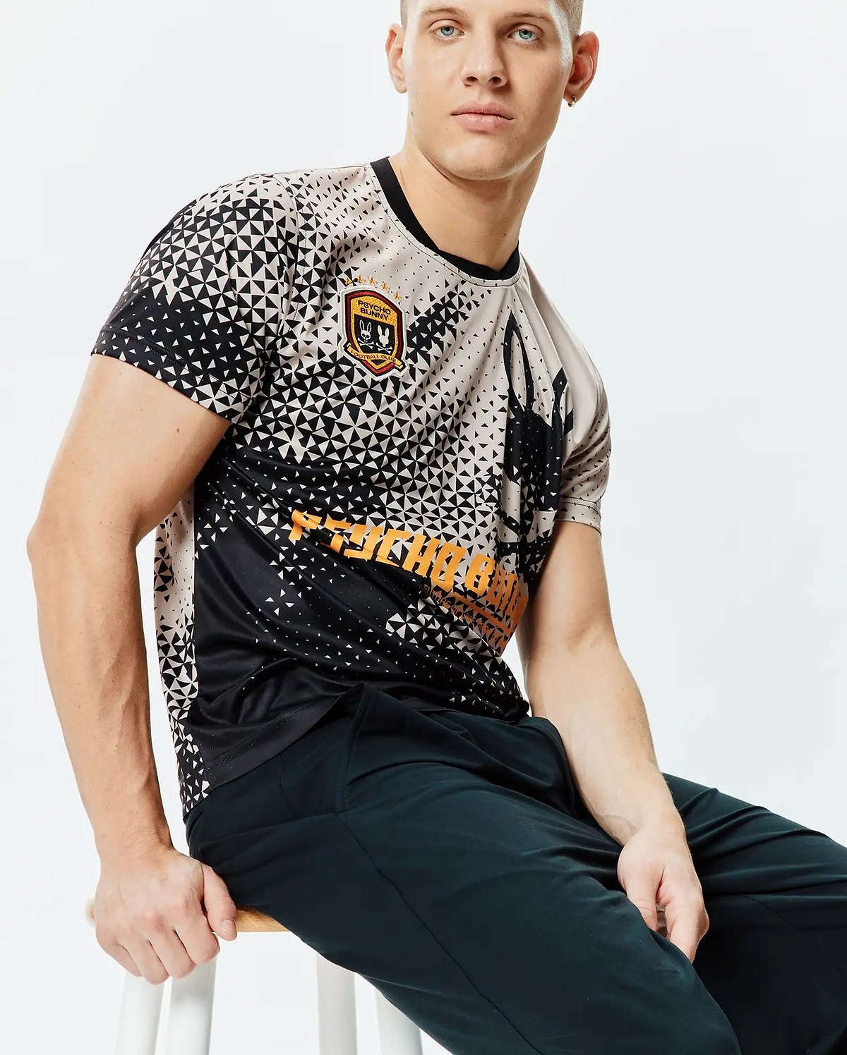 Mens Joshua Soccer Tee, a classic fit black and beige geometric triangle pattern jersey with an embroidered Psycho Bunny Football Club patch, large bunny skull graphic, and bold orange Psycho Bunny text. (3)
