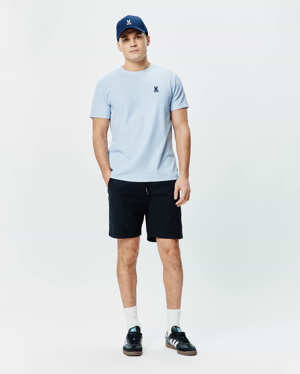 Light blue Mens Taylor Pique Tee with a classic fit and small embroidered Psycho Bunny skull logo on the left chest, paired with a navy cap and black shorts.