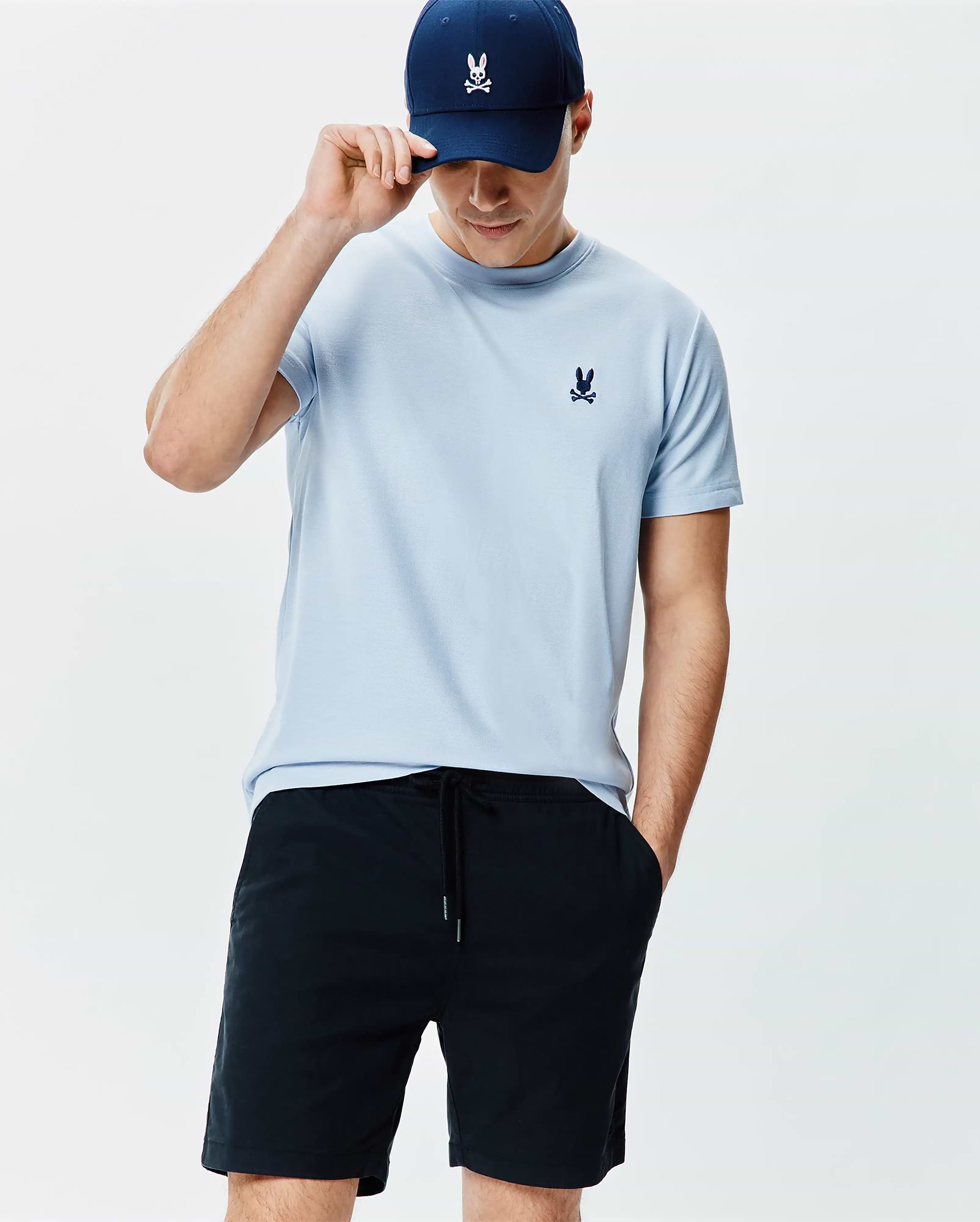 Mens Taylor Pique Tee in light blue pique knit, featuring a small navy embroidered Psycho Bunny logo on the chest, paired with navy drawstring shorts and a navy baseball cap with a white bunny skull.