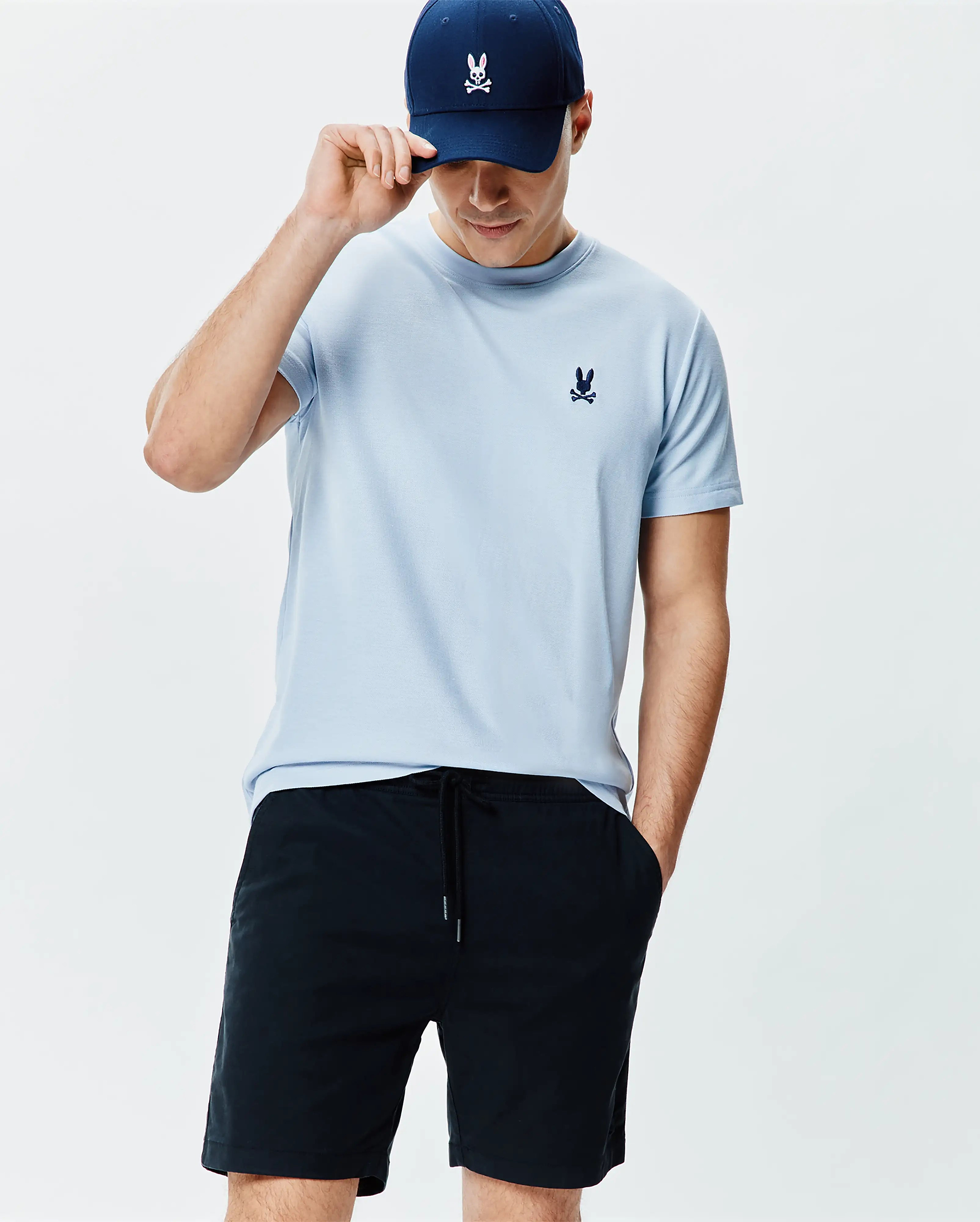 Mens Taylor Pique Tee in light blue pique knit, featuring a small navy embroidered Psycho Bunny logo on the chest, paired with navy drawstring shorts and a navy baseball cap with a white bunny skull.