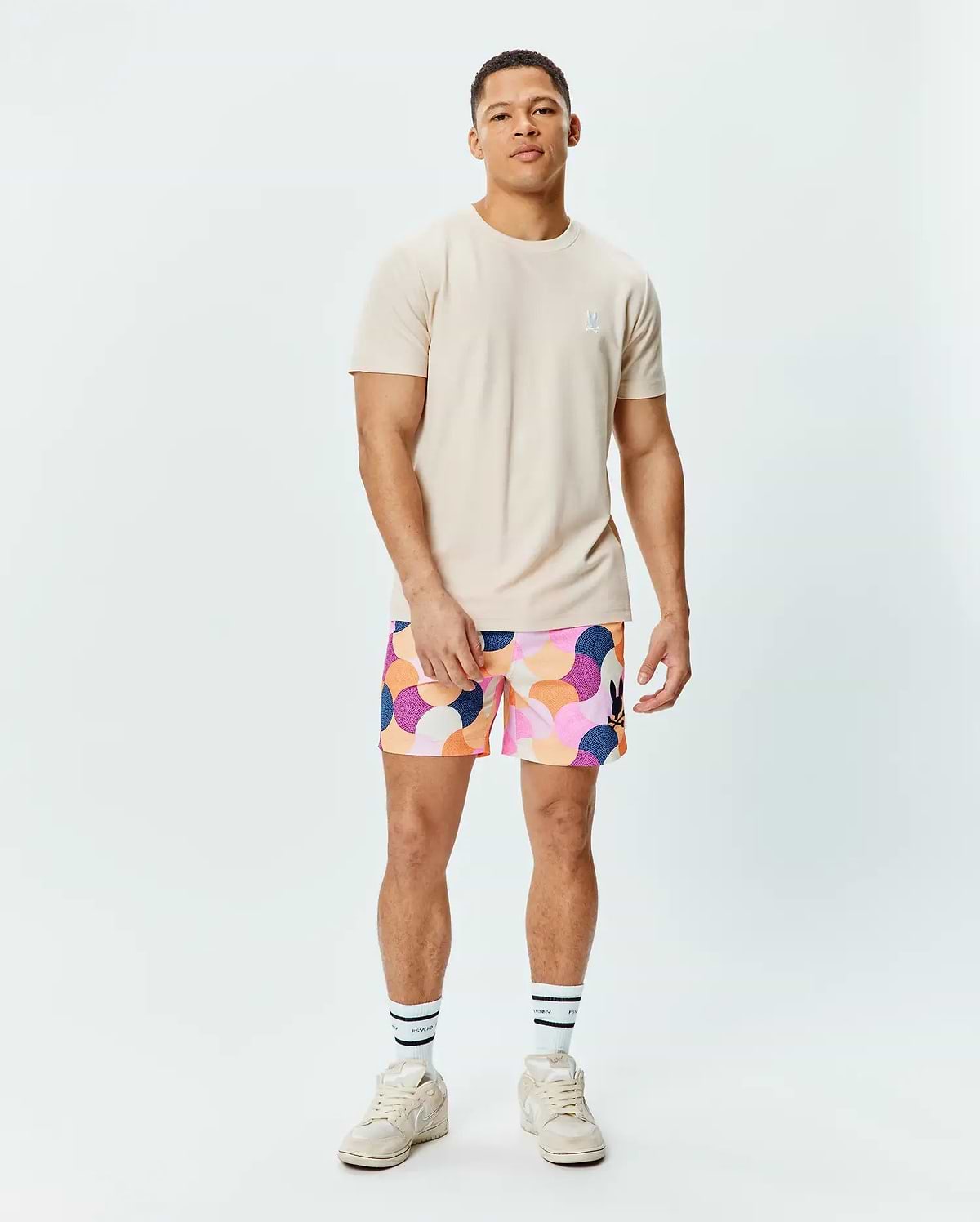 Mens Taylor Pique Tee in light beige with a small embroidered white Psycho Bunny logo on the left chest, paired with multi-color abstract patterned shorts featuring a black bunny logo on the right thigh.