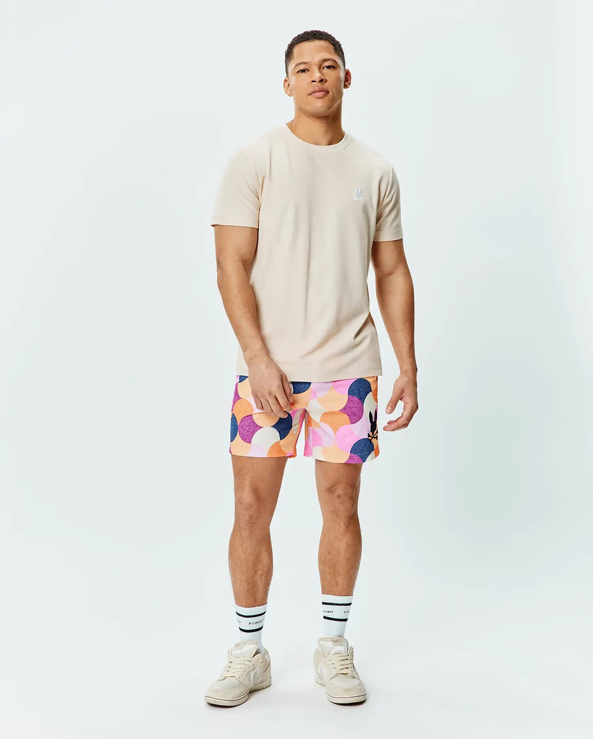 Mens Taylor Pique Tee in light beige with a small embroidered white Psycho Bunny logo on the left chest, paired with multi-color abstract patterned shorts featuring a black bunny logo on the right thigh.