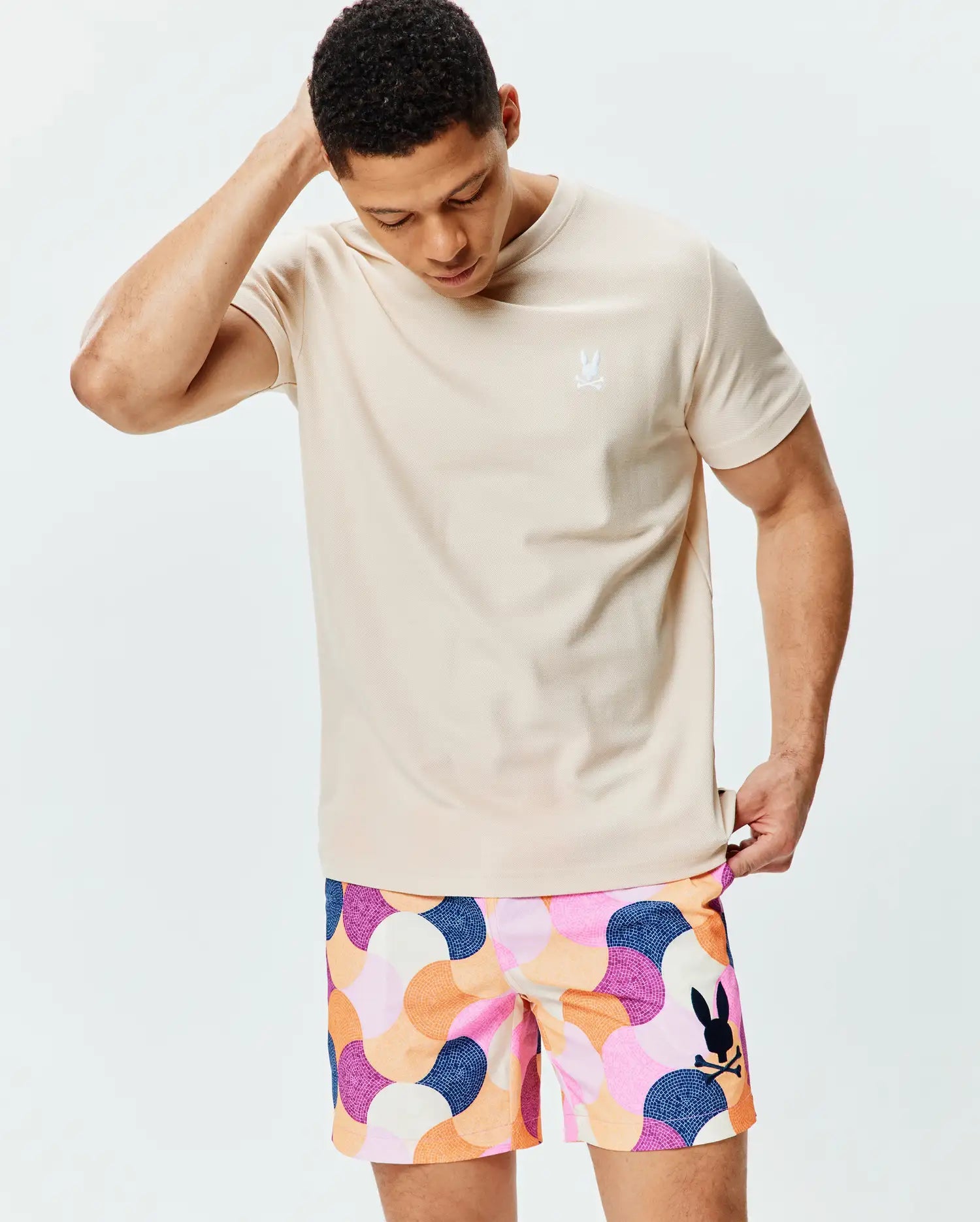 Mens Taylor Pique Tee in light beige pique knit with a small white embroidered Psycho Bunny logo on the left chest, paired with multi-colored geometric mosaic print shorts featuring a large black Psycho Bunny logo.
