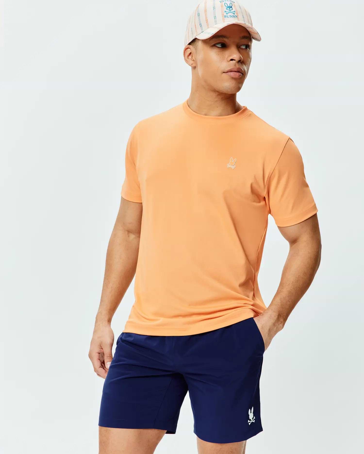 Mens Dash Bunny Sport Tee in peach jersey with a small embroidered white Psycho Bunny logo on the chest, paired with navy shorts featuring a white bunny skull, and a striped cap.