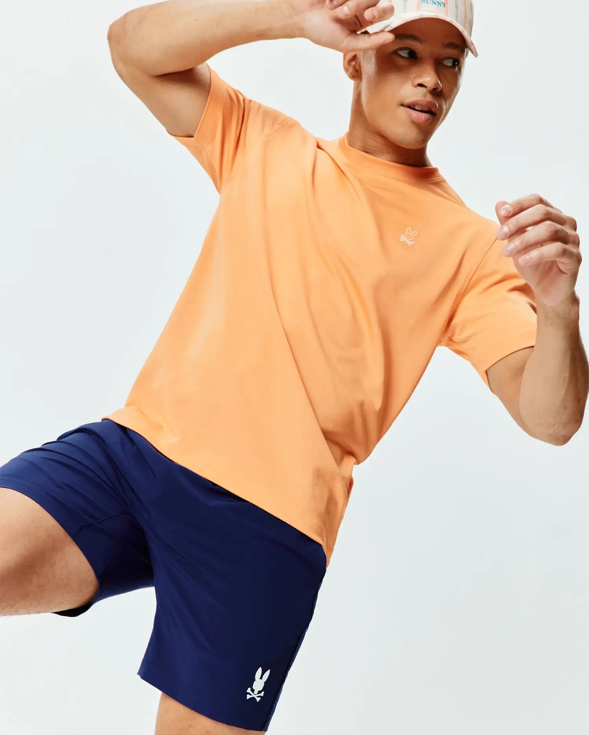 Mens Dash Bunny Sport Tee. Peach, relaxed fit, smooth jersey tee with a small embroidered white bunny skull logo on the left chest, complemented by navy performance shorts with a white bunny skull logo.