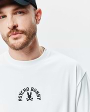 White Mens Stevie Relaxed Fit Tee with a smooth jersey texture, featuring a black embroidered Psycho Bunny skull and crossbones logo with arched text on the chest.