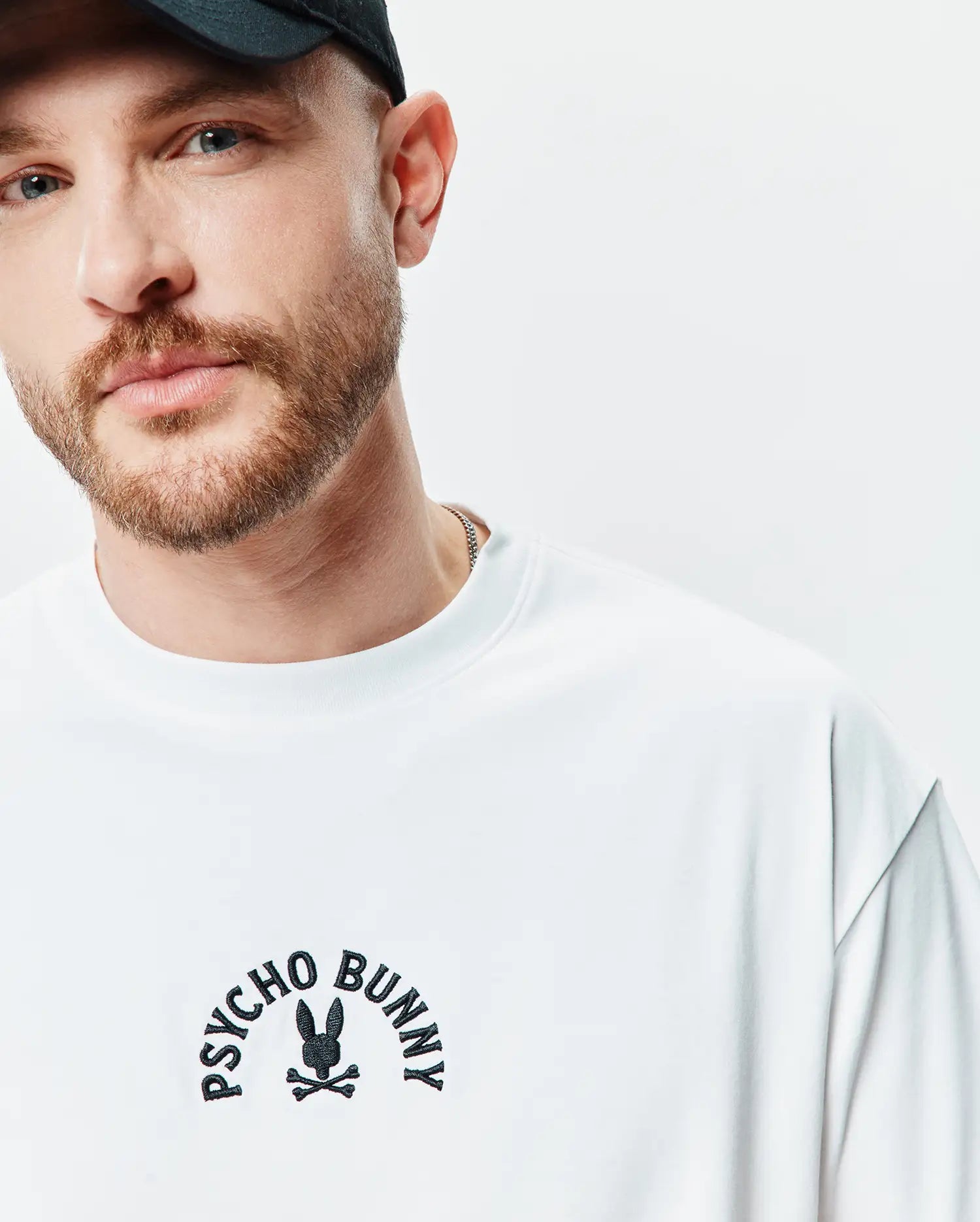 White Mens Stevie Relaxed Fit Tee with a smooth jersey texture, featuring a black embroidered Psycho Bunny skull and crossbones logo with arched text on the chest.