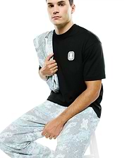 Men's Samson Oversized Bunny Tee: Black ribbed t-shirt with an oversized fit, featuring a small white embroidered Psycho Bunny skull logo on the left chest. Complemented by light blue digital camo pants. (3)