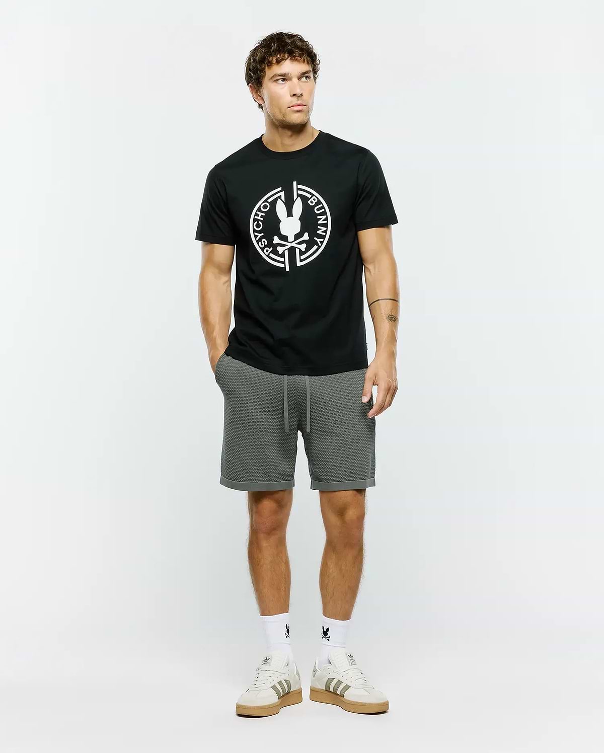 Mens Santa Fe Bunny Graphic Tee, a black classic fit premium jersey t-shirt featuring a large white circular Psycho Bunny skull logo graphic on the chest, complemented by grey textured knit drawstring shorts with a cuffed hem.