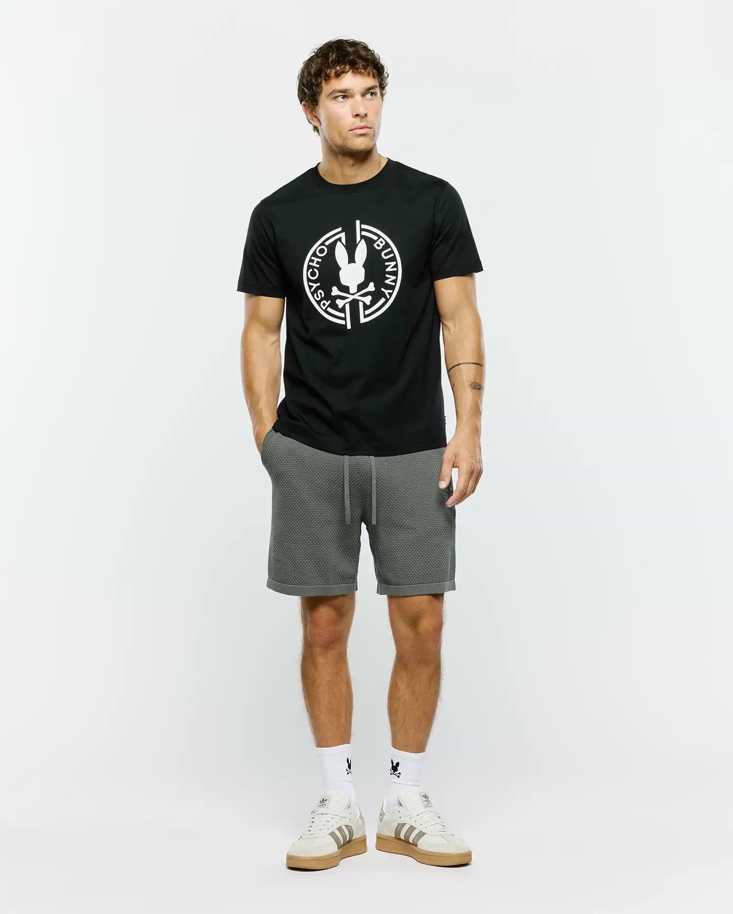 Mens Santa Fe Bunny Graphic Tee, a black classic fit premium jersey t-shirt featuring a large white circular Psycho Bunny skull logo graphic on the chest, complemented by grey textured knit drawstring shorts with a cuffed hem.
