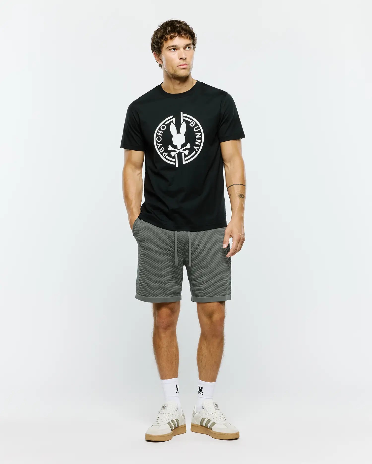 Mens Santa Fe Bunny Graphic Tee, a black classic fit premium jersey t-shirt featuring a large white circular Psycho Bunny skull logo graphic on the chest, complemented by grey textured knit drawstring shorts with a cuffed hem.