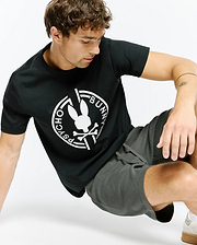 Mens Santa Fe Bunny Graphic Tee, a black classic-fit jersey t-shirt featuring a large white circular Psycho Bunny skull and crossbones graphic on the chest, styled with dark grey waffle knit drawstring shorts.