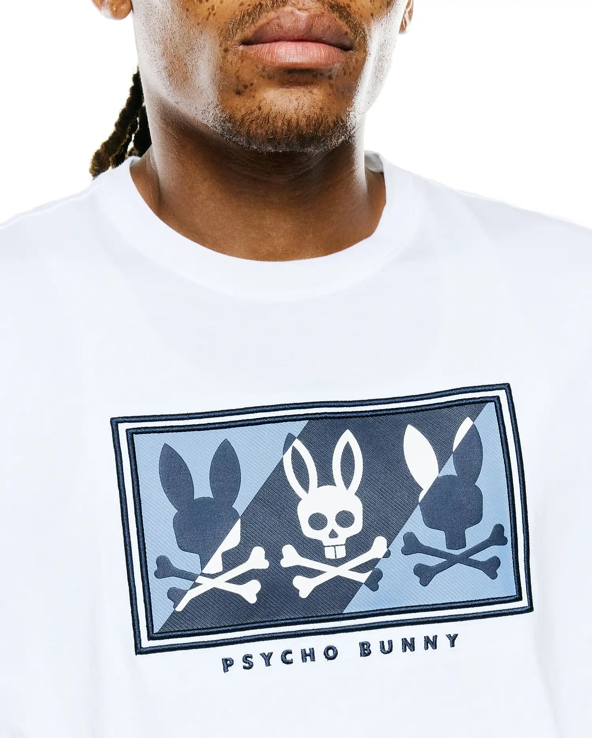 Mens Desmond Graphic Tee in white jersey knit, featuring a large chest graphic with three bunny skull and silhouette designs in white, light blue, and dark blue on a patterned background, bordered in dark blue, with PSYCHO BUNNY text below.