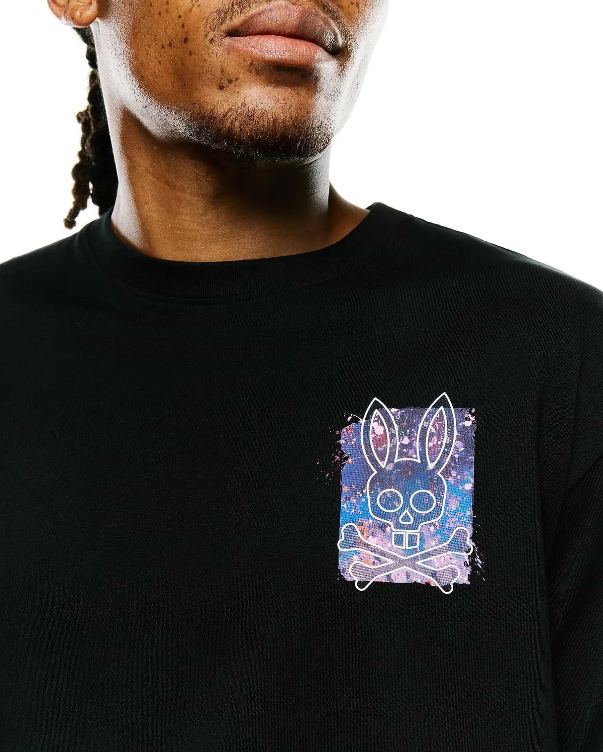 Mens Isaac Splatter Back Graphic Tee, a black jersey t-shirt with a small left chest graphic featuring a white Psycho Bunny skull and crossbones over a vibrant purple, blue, and pink splatter background.