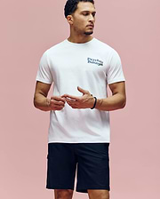 A man stands against a pink background, wearing the Psycho Bunny MENS ARCHIVE WORDMARK GRAPHIC TEE (MT0200030) in white Pima cotton and black shorts. He looks to the side with his hands loosely clasped in front of him. (2)