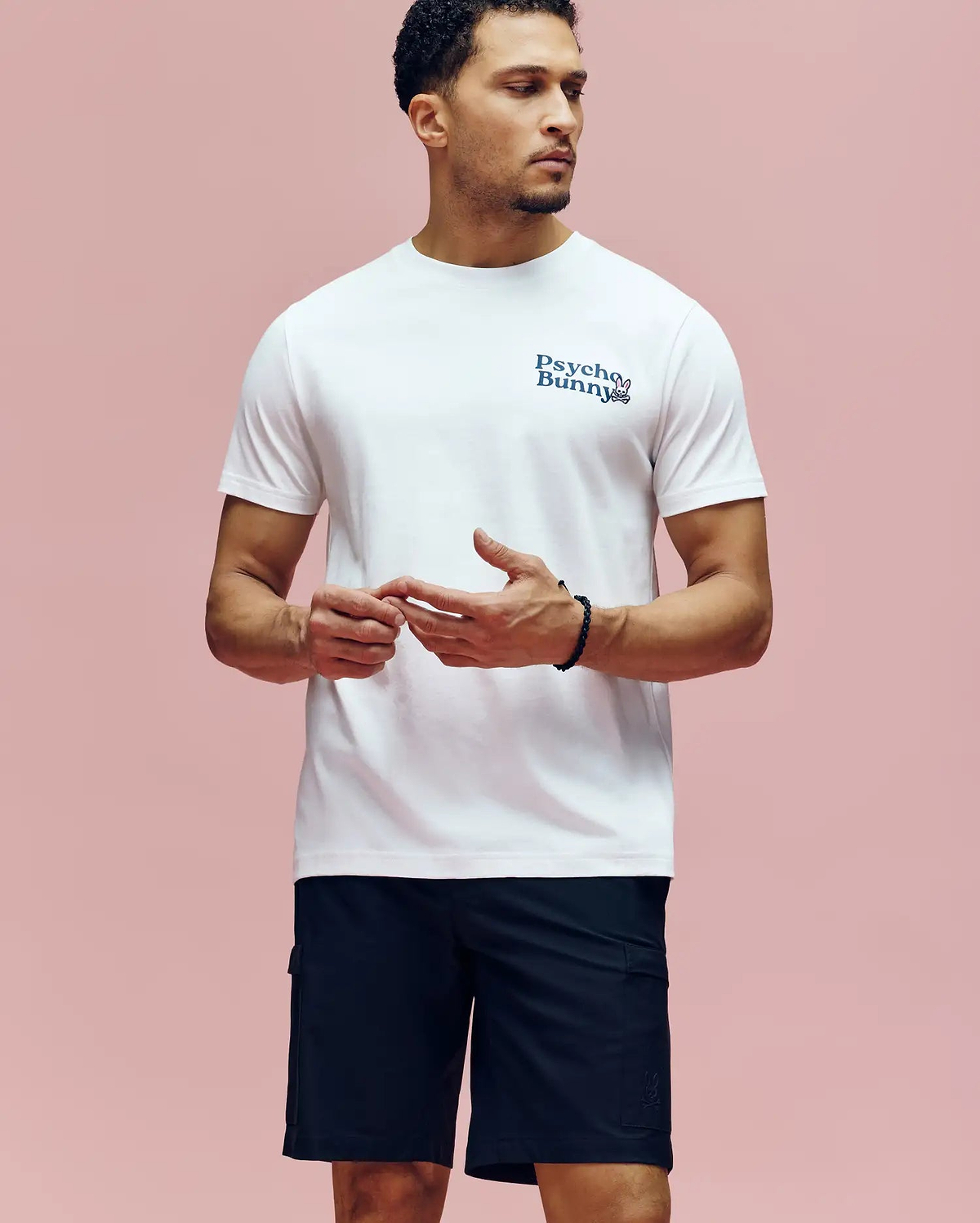 A man stands against a pink background, wearing the Psycho Bunny MENS ARCHIVE WORDMARK GRAPHIC TEE (MT0200030) in white Pima cotton and black shorts. He looks to the side with his hands loosely clasped in front of him. (2)