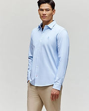 A man in the Psycho Bunny MENS JIM JERSEY STRIPED KNIT SHIRT (MT0300041), light blue, stands slightly turned with a neutral expression against a plain light background, paired with beige pants.