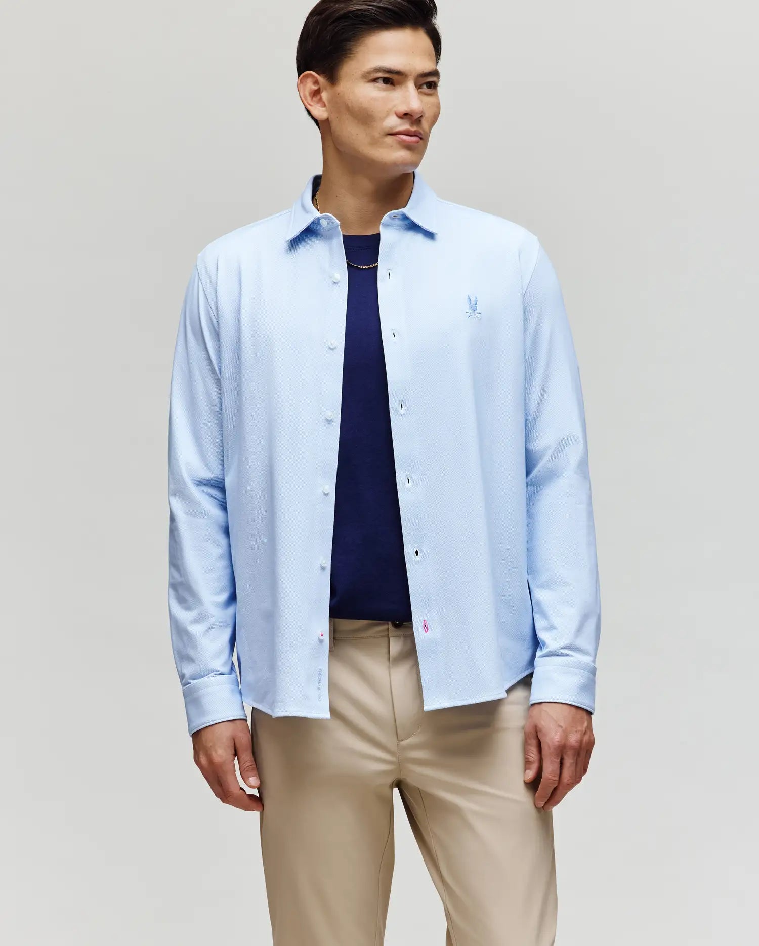A man stands slightly angled against a plain light background, wearing the Psycho Bunny MENS JIM JERSEY STRIPED KNIT SHIRT (MT0300041) over a navy cotton jersey tee and beige pants.