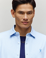 A man with short dark hair wears the Psycho Bunny MENS JIM JERSEY STRIPED KNIT SHIRT (MT0300041) over a dark shirt and gold chain, standing against a plain light background.