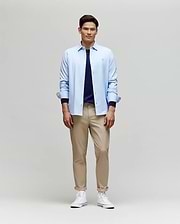 A man smiles while facing forward, wearing the Psycho Bunny MENS JIM JERSEY STRIPED KNIT SHIRT (MT0300041) over a navy tee, paired with beige pants and white high-top sneakers against a plain light background.