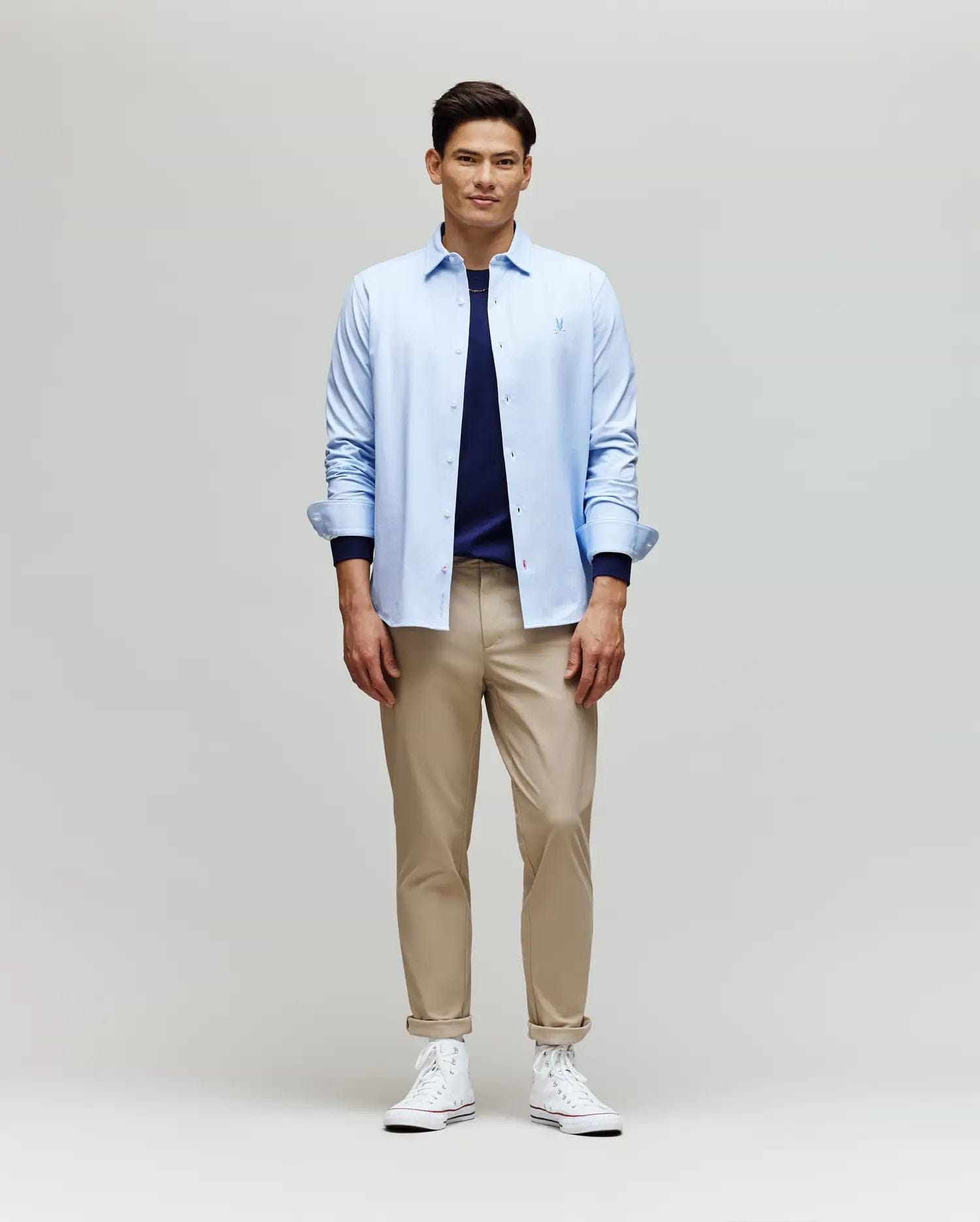 A man smiles while facing forward, wearing the Psycho Bunny MENS JIM JERSEY STRIPED KNIT SHIRT (MT0300041) over a navy tee, paired with beige pants and white high-top sneakers against a plain light background.