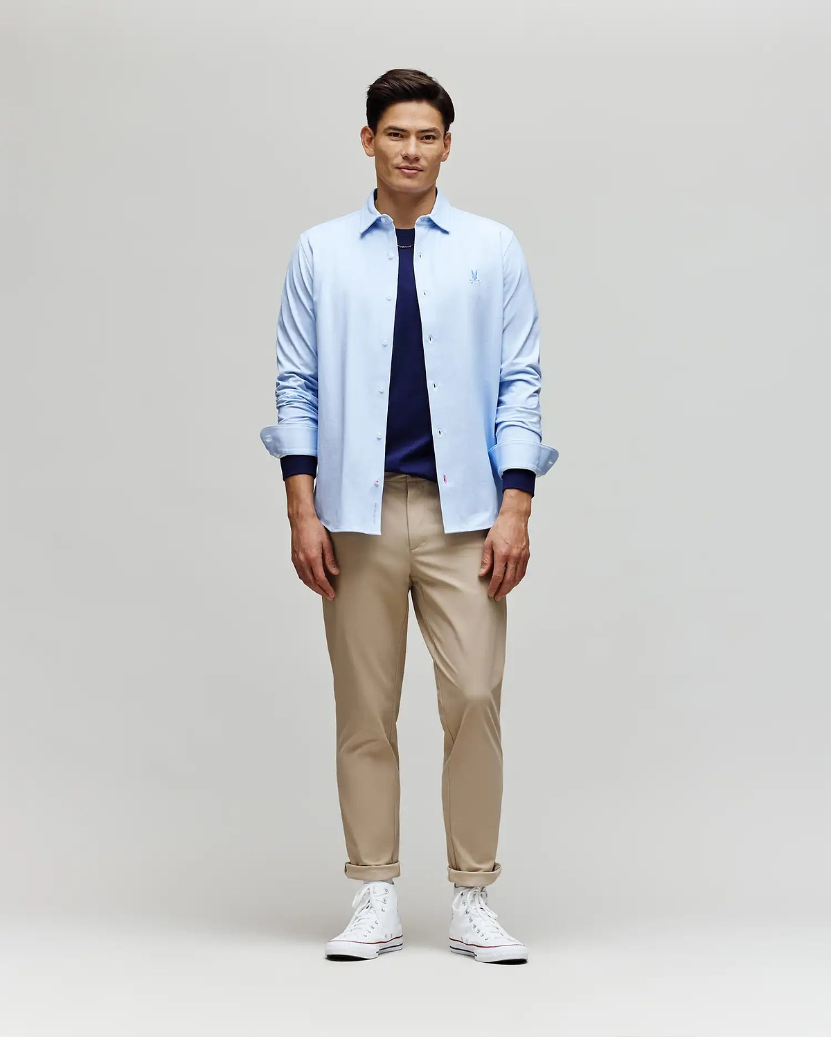 A man smiles while facing forward, wearing the Psycho Bunny MENS JIM JERSEY STRIPED KNIT SHIRT (MT0300041) over a navy tee, paired with beige pants and white high-top sneakers against a plain light background.