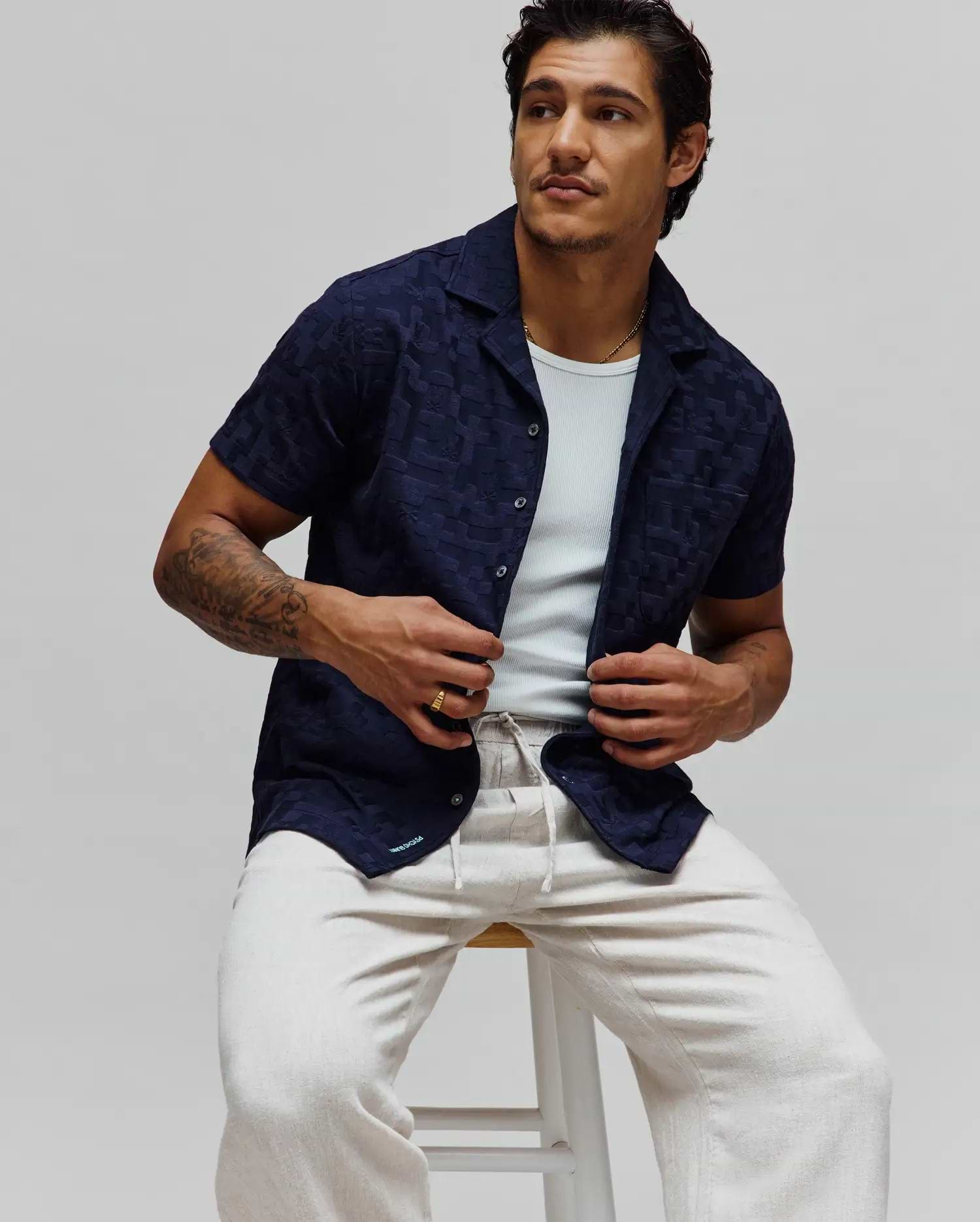 Navy blue Mens Josh Jacquard Shirt featuring a subtle tonal geometric pattern, relaxed fit, short sleeves, camp collar, and a small light blue Psycho Bunny logo on the bottom hem.