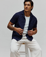 Navy blue Mens Josh Jacquard Shirt featuring a subtle tonal geometric pattern, relaxed fit, short sleeves, camp collar, and a small light blue Psycho Bunny logo on the bottom hem.