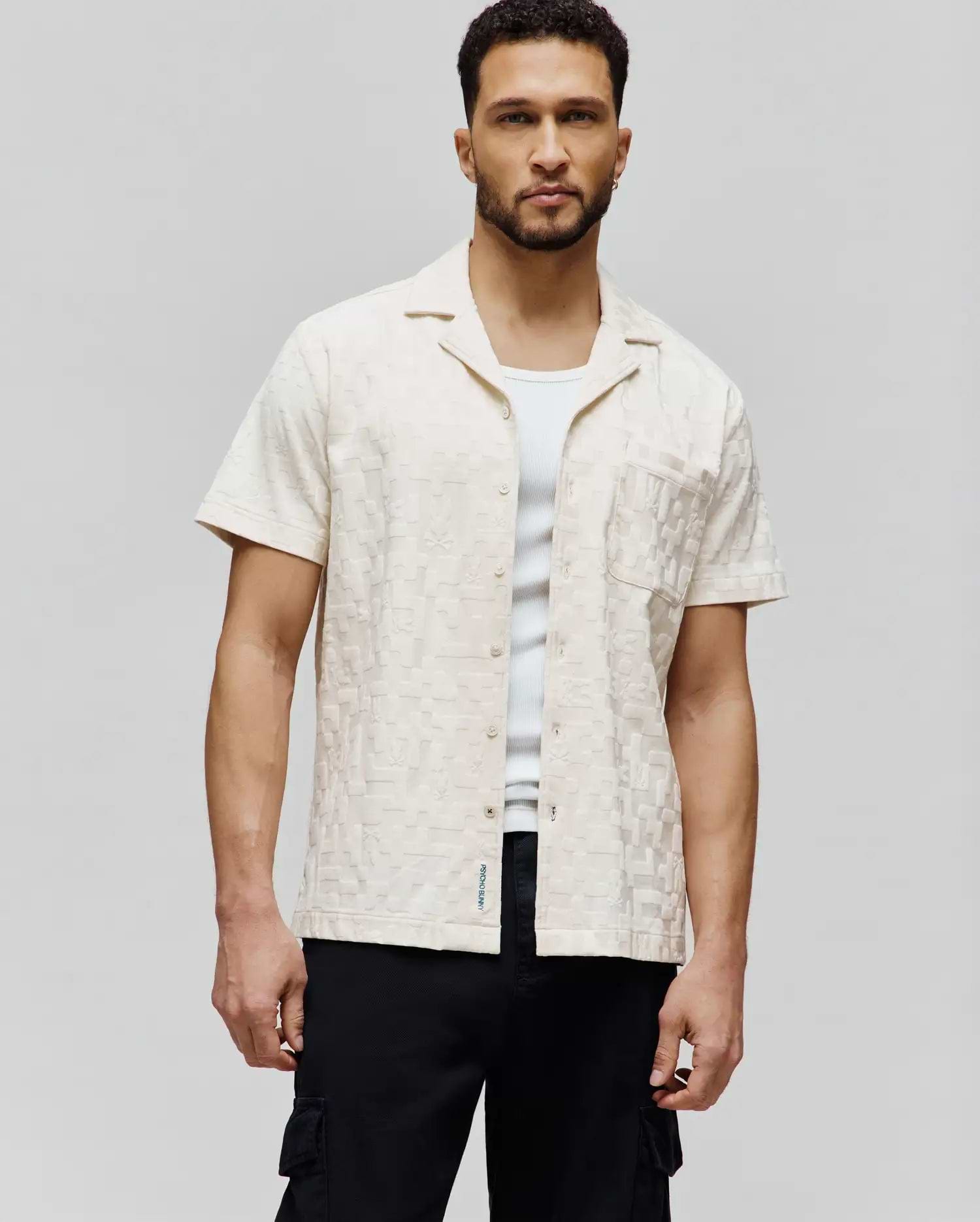Mens Josh Jacquard Shirt, a cream short-sleeve shirt with all-over tonal bunny skull jacquard pattern, classic fit, chest pocket, and small blue Psycho Bunny text logo on the hem.
