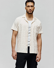 Mens Josh Jacquard Shirt, a cream short-sleeve shirt with all-over tonal bunny skull jacquard pattern, classic fit, chest pocket, and small blue Psycho Bunny text logo on the hem.
