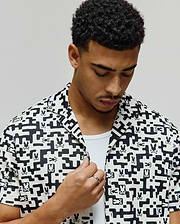 Mens Josh Jacquard Shirt, a white short-sleeve camp collar shirt with a black geometric pixelated jacquard pattern featuring integrated small Psycho Bunny skull logos, classic fit.