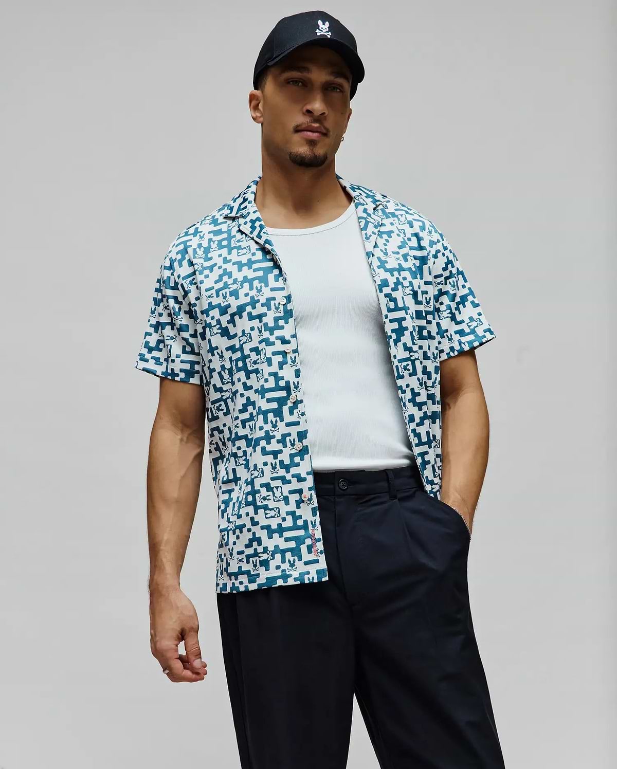 Mens Josh Jacquard Shirt, a relaxed fit short-sleeve camp collar shirt in white with an all-over teal pixelated Psycho Bunny skull pattern and a red side tag.