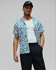 Mens Josh Jacquard Shirt, a relaxed fit short-sleeve camp collar shirt in white with an all-over teal pixelated Psycho Bunny skull pattern and a red side tag.