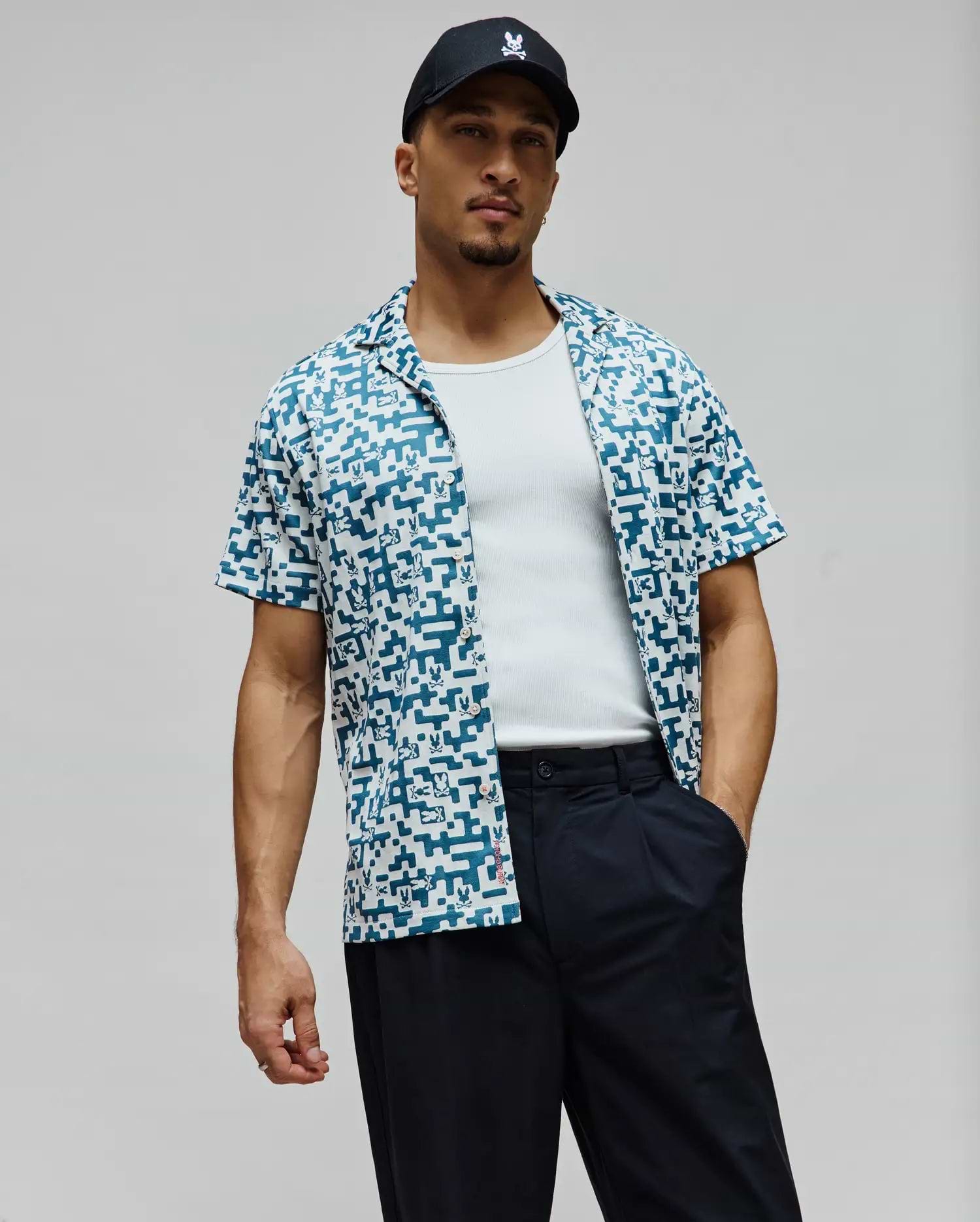 Mens Josh Jacquard Shirt, a relaxed fit short-sleeve camp collar shirt in white with an all-over teal pixelated Psycho Bunny skull pattern and a red side tag.