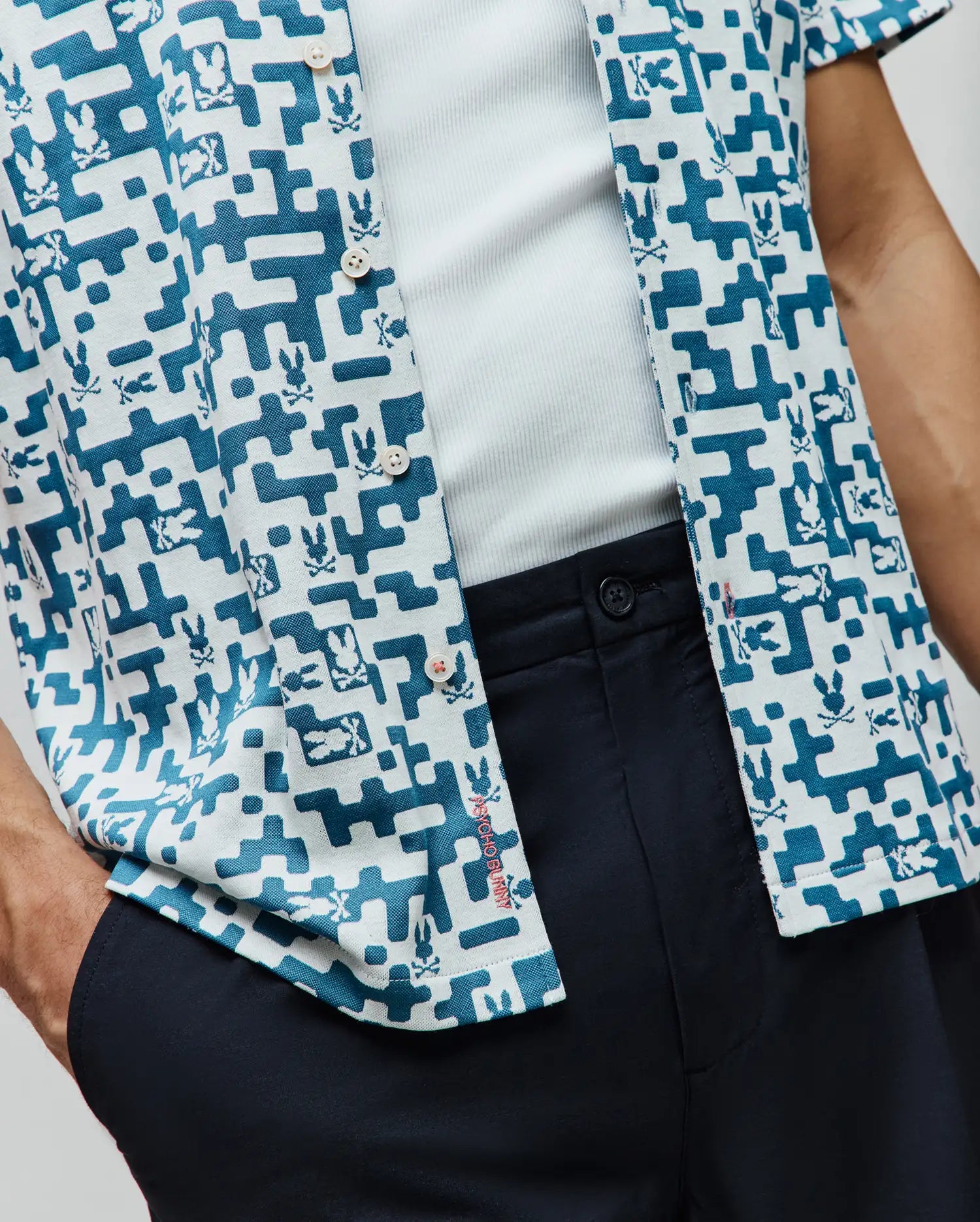 Mens Josh Jacquard Shirt, a relaxed fit short-sleeve button-up featuring a white and teal pixelated geometric pattern with repeated Psycho Bunny skull logos and subtle red placket branding.