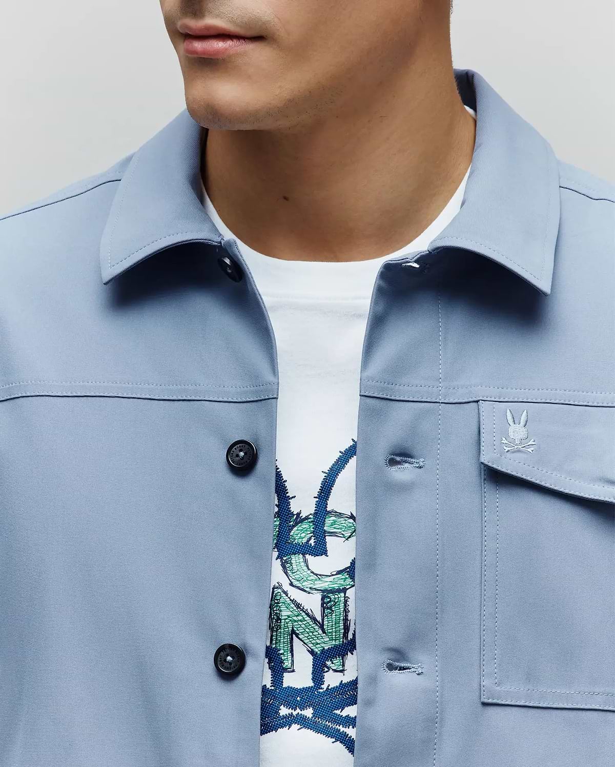 Mens Maverick Overshirt in periwinkle blue, a classic fit with smooth fabric, black buttons, and a small embroidered Psycho Bunny skull logo on the left chest pocket. A white graphic tee is visible underneath. (2)
