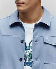 Mens Maverick Overshirt in periwinkle blue, a classic fit with smooth fabric, black buttons, and a small embroidered Psycho Bunny skull logo on the left chest pocket. A white graphic tee is visible underneath. (2)