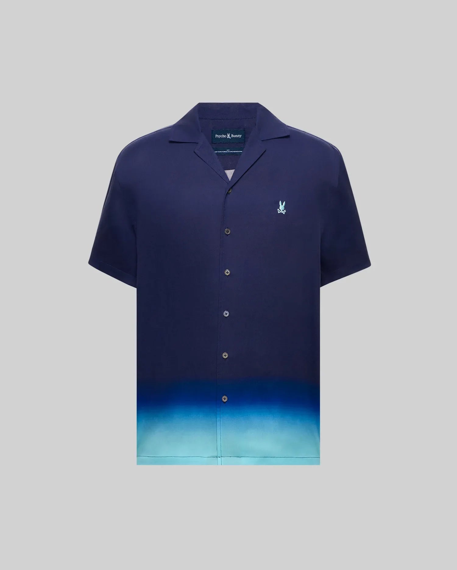MENS DAX TENCEL SHIRT, a relaxed fit navy blue short-sleeve camp collar shirt with an ombre gradient fading to light blue at the hem and a small embroidered Psycho Bunny skull logo on the left chest.