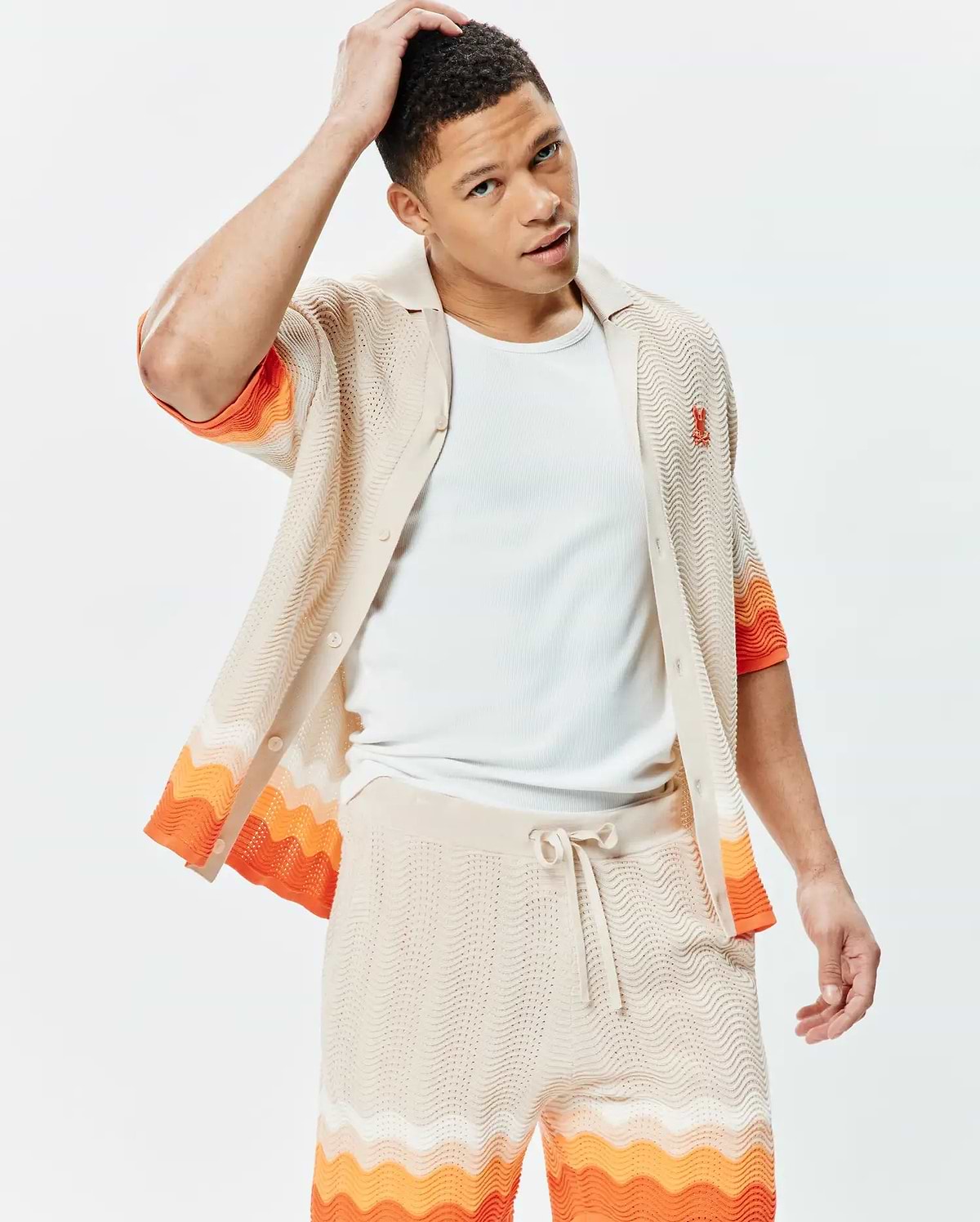 Mens Holden Relaxed Fit Knit Shirt in beige with a wavy knit texture, featuring vibrant orange and white horizontal wavy stripes on sleeves and hem, and a small embroidered orange Psycho Bunny skull logo on the left chest, paired with matching knit shorts.