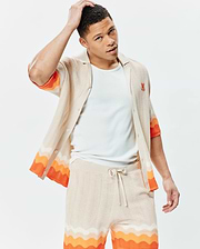 Mens Holden Relaxed Fit Knit Shirt in beige with a wavy knit texture, featuring vibrant orange and white horizontal wavy stripes on sleeves and hem, and a small embroidered orange Psycho Bunny skull logo on the left chest, paired with matching knit shorts.