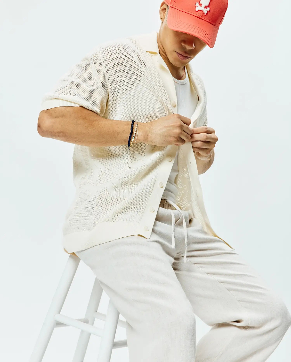 Mens Brad Oversized Knit Shirt in cream, featuring an open-knit texture, short sleeves, and a collared button-down front, paired with light beige drawstring pants and a coral Psycho Bunny logo cap.