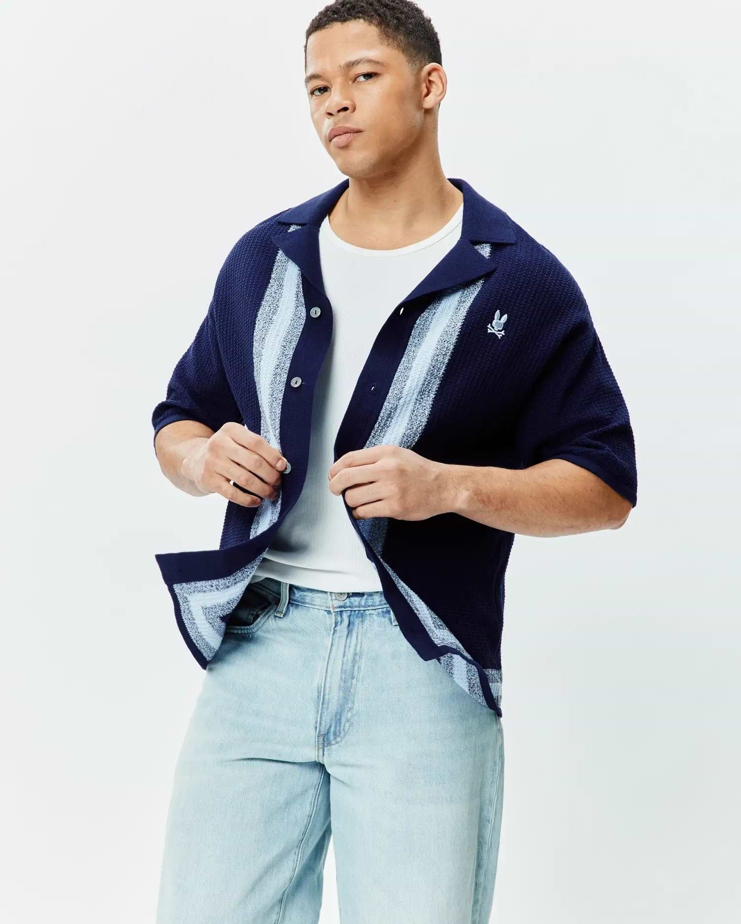 Mens Giles Relaxed Fit Border Knit Shirt in navy blue textured knit, featuring light blue and white vertical stripes on the placket and hem, with a small embroidered Psycho Bunny skull logo on the left chest.