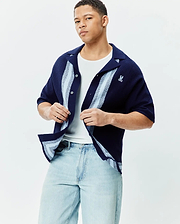 Mens Giles Relaxed Fit Border Knit Shirt in navy blue textured knit, featuring light blue and white vertical stripes on the placket and hem, with a small embroidered Psycho Bunny skull logo on the left chest.