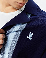 Mens Giles Relaxed Fit Border Knit Shirt in navy blue textured knit, featuring light blue and white vertical stripes, a spread collar, and a small embroidered Psycho Bunny skull logo on the left chest.