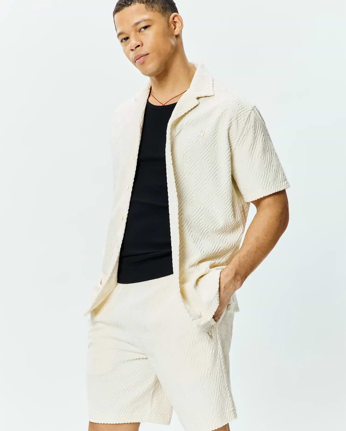 Mens Tristan Shirt: Relaxed fit cream short-sleeve shirt with embossed geometric diamond pattern, textured knit fabric, and small embroidered Psycho Bunny skull logo on left chest, paired with matching shorts.