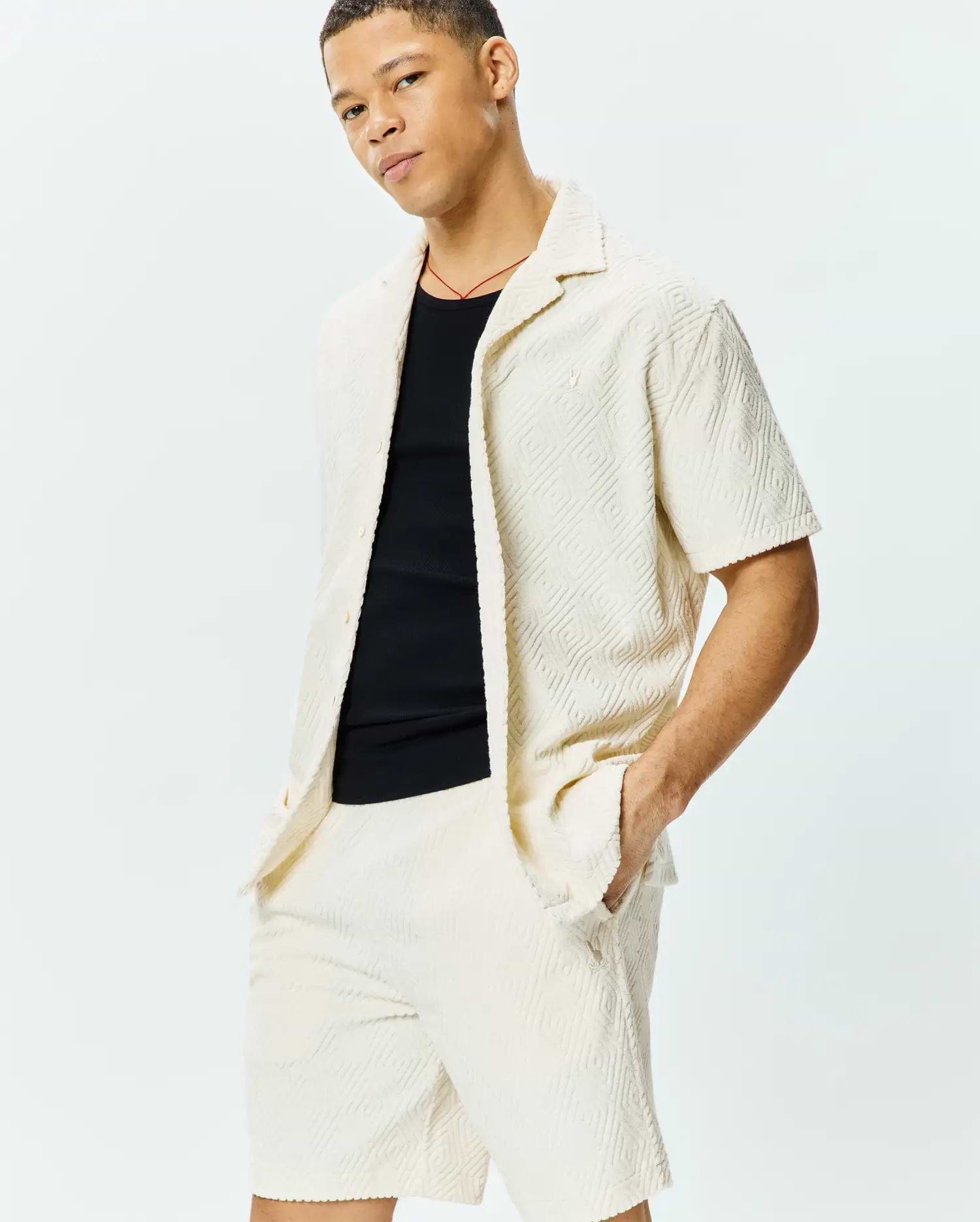 Mens Tristan Shirt: Relaxed fit cream short-sleeve shirt with embossed geometric diamond pattern, textured knit fabric, and small embroidered Psycho Bunny skull logo on left chest, paired with matching shorts.
